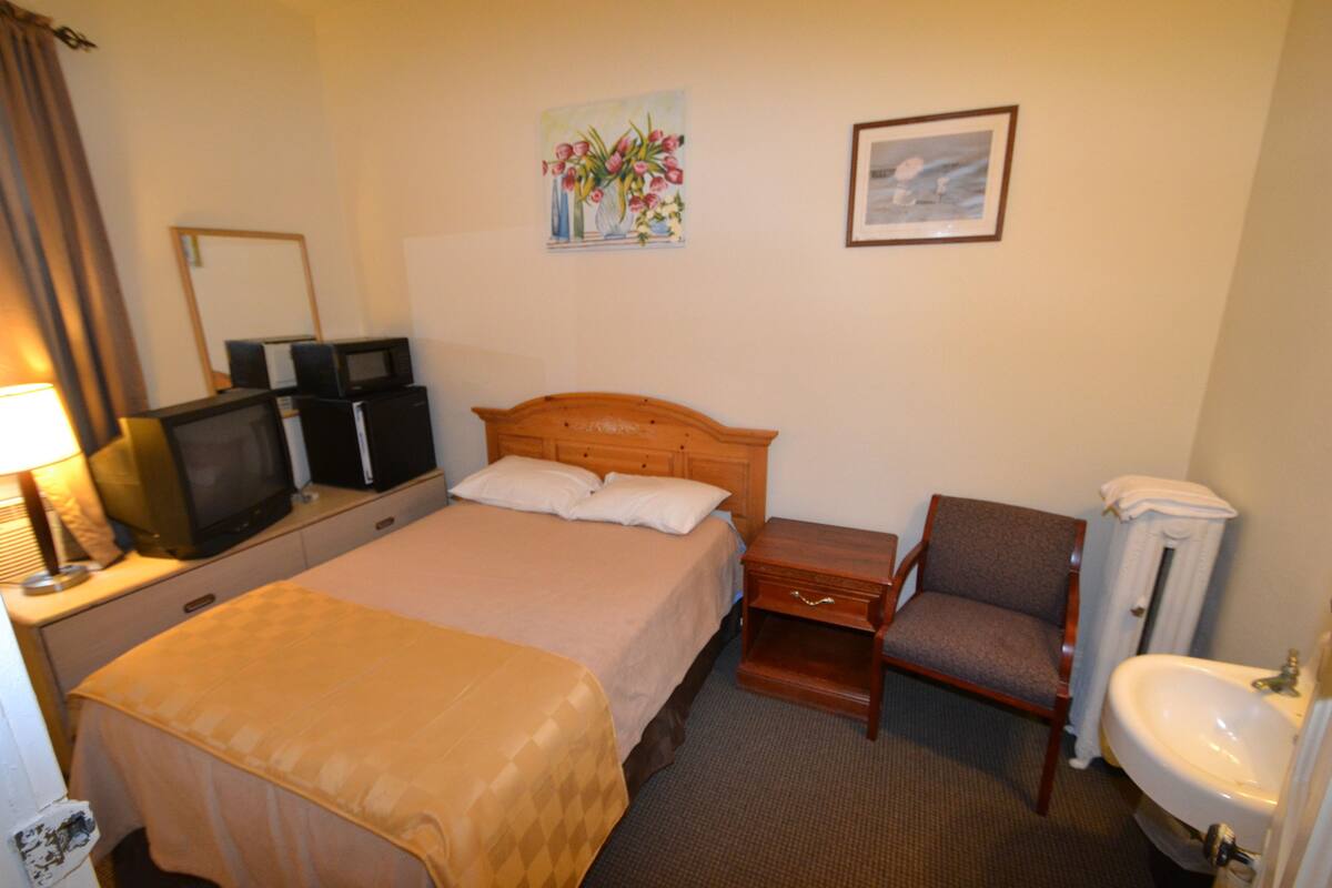 A hotel room with a bed, a chair, a desk with a television, a mirror, a painting, and a sink.