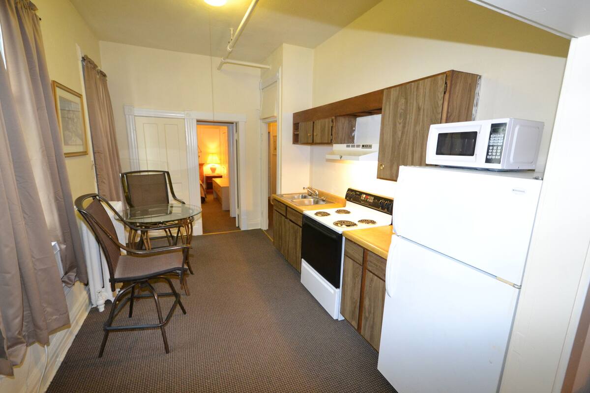 A compact kitchen with a stove, microwave, and refrigerator. There is a dining table with chairs and a small island. The room has a carpeted floor and wooden cabinets.