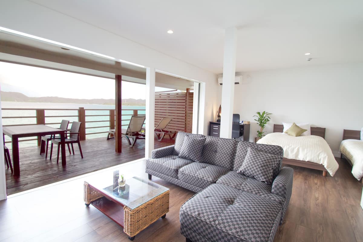 A modern hotel room with a large sofa, a bed, a dining area with chairs, and a view of the sea.