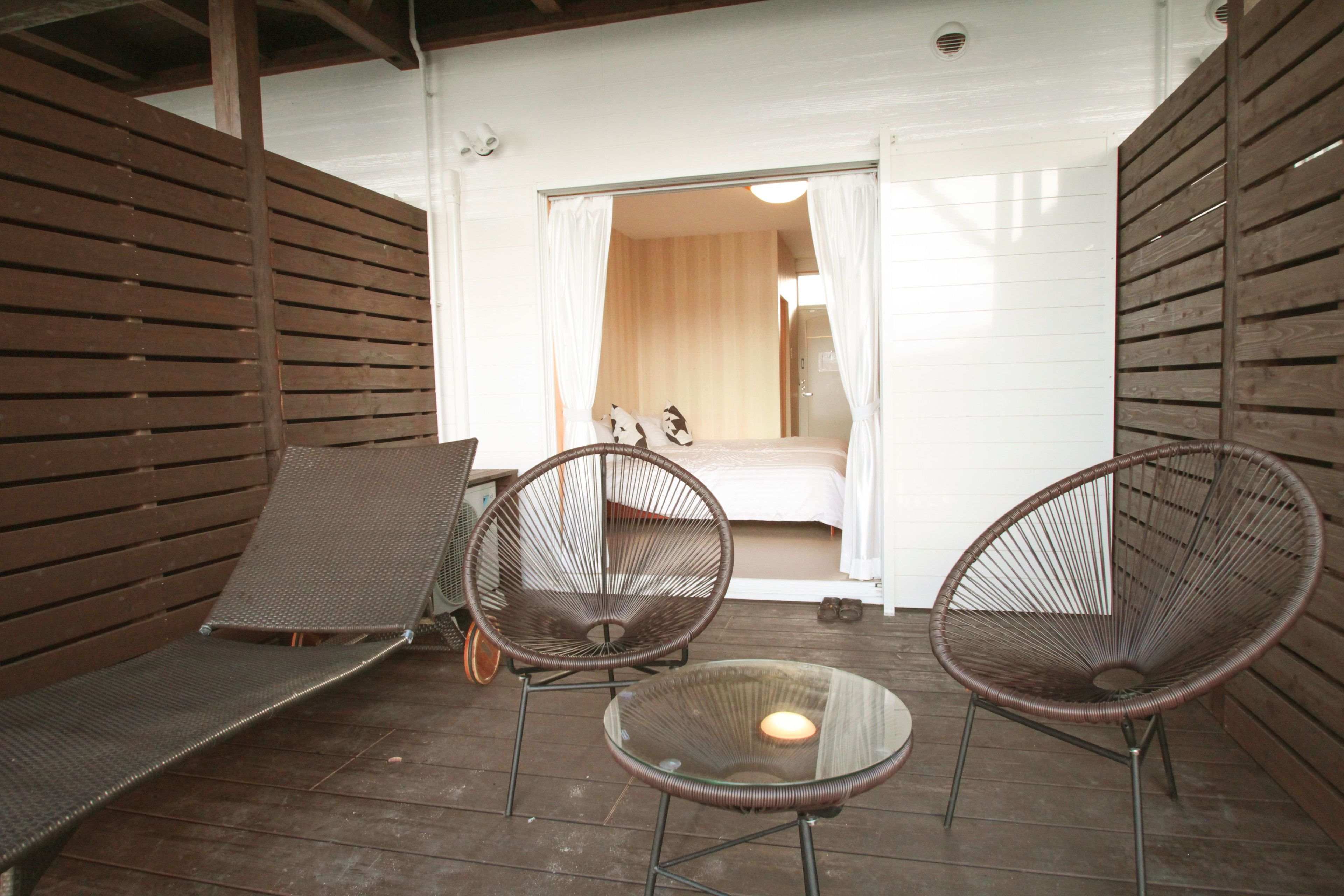 An outdoor seating area with wicker chairs and a glass table, a wooden deck, and a doorway leading to a room with a bed and curtains.