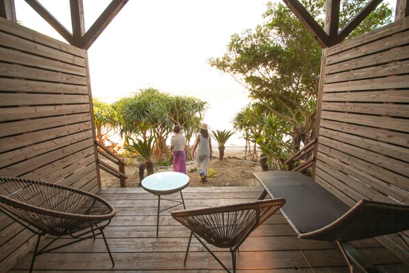 A wooden deck with wicker chairs and a small table, overlooking a beach with palm trees and a person walking. - Native Sea Amami Adan On The Beach (Tatsugo)