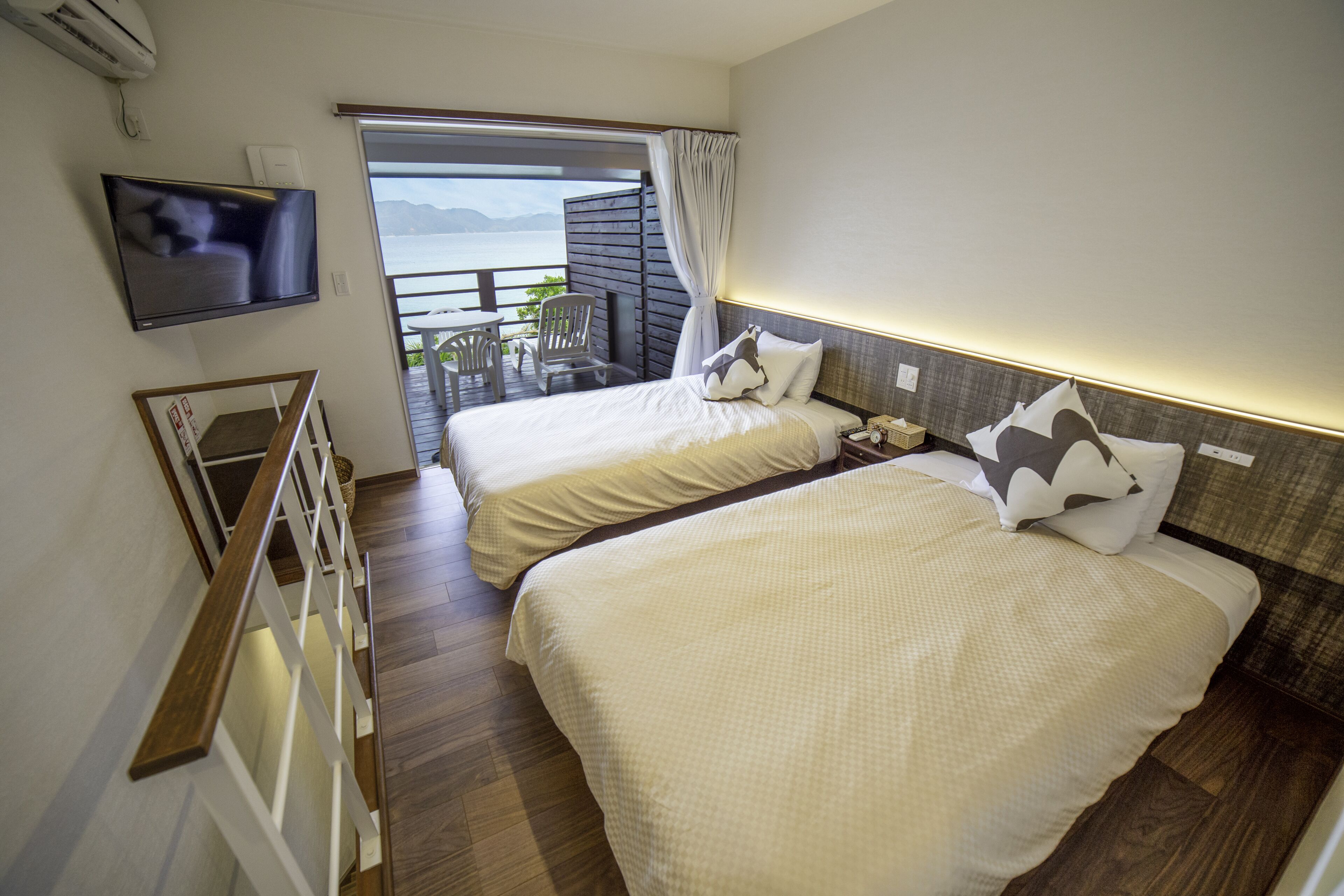 Habitación de hotel con dos camas, un televisor montado en la pared, un balcón con mesa y sillas, y vista al mar.