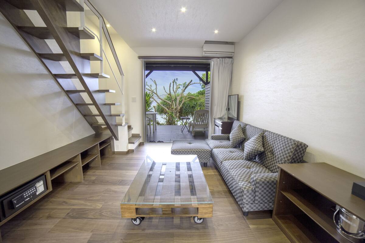 A modern living room with a glass coffee table, a patterned sofa, a staircase, and a view through a glass door.