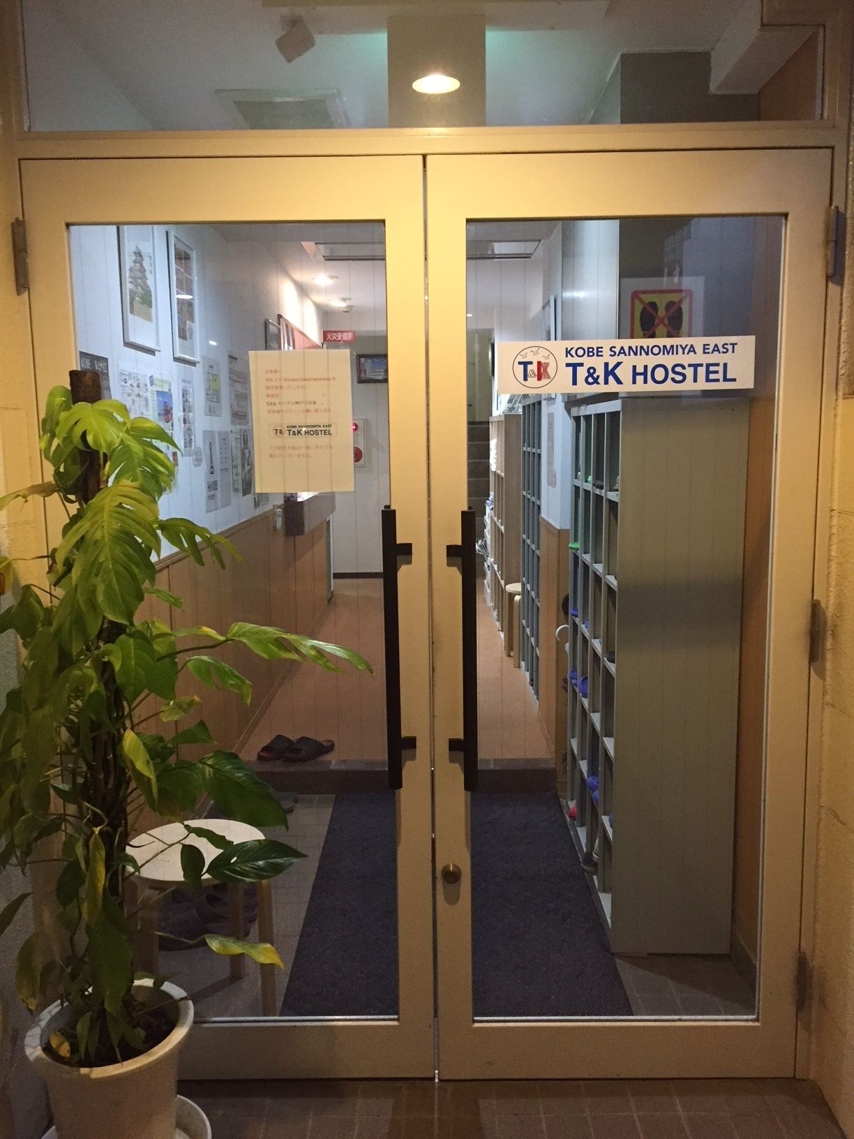 A T&K Hostel entrance with a potted plant, a sign indicating it's the Kobe Sannomiya East location, and a hallway leading to rooms.