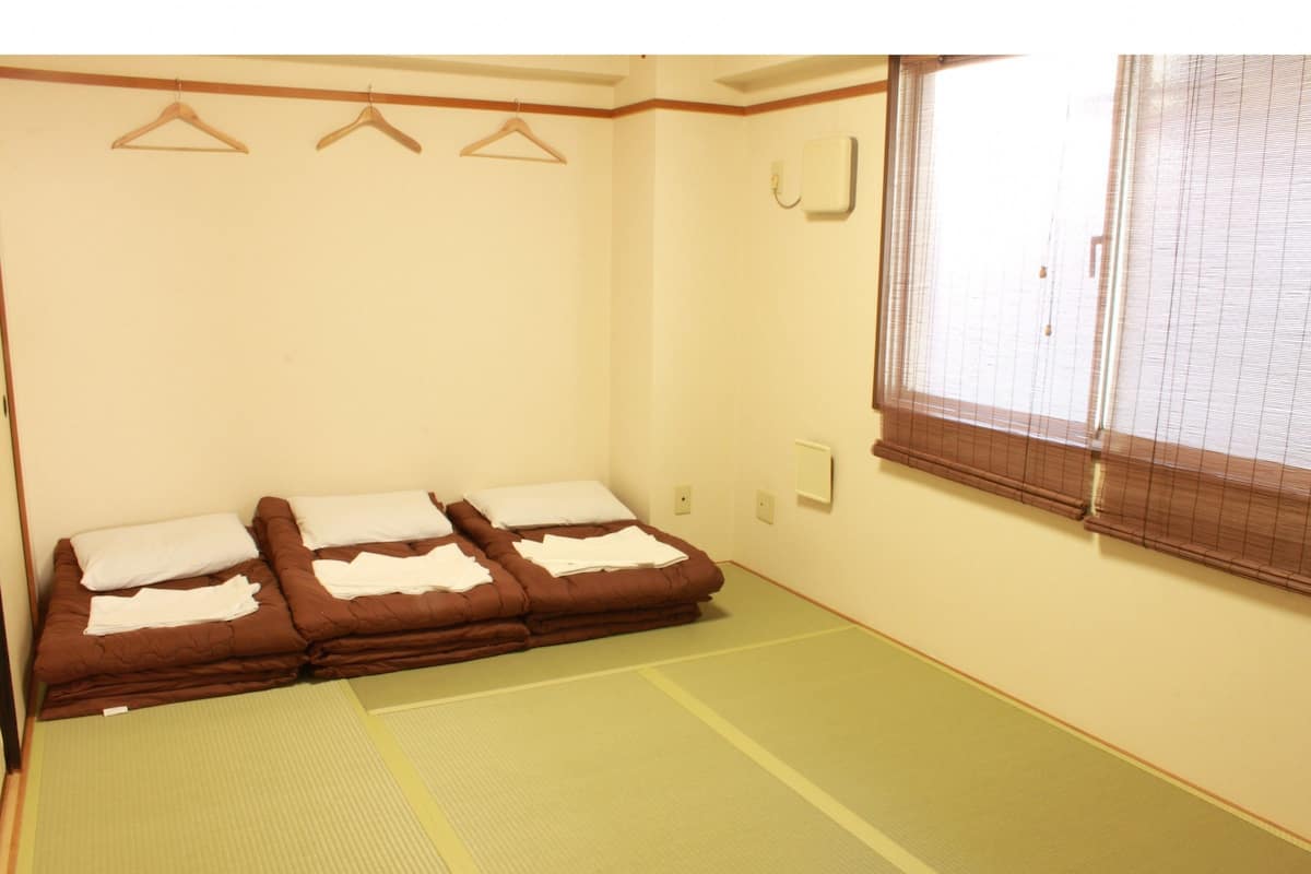 Ein Zimmer mit Tatami-Böden, zwei Betten mit weißer und brauner Bettwäsche, Holzaufhänger an der Wand und einem Fenster mit Jalousien.