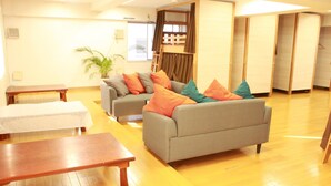 A living room with a grey sofa, wooden coffee table, and a small table with a white tablecloth. There is a potted plant and a window with curtains.