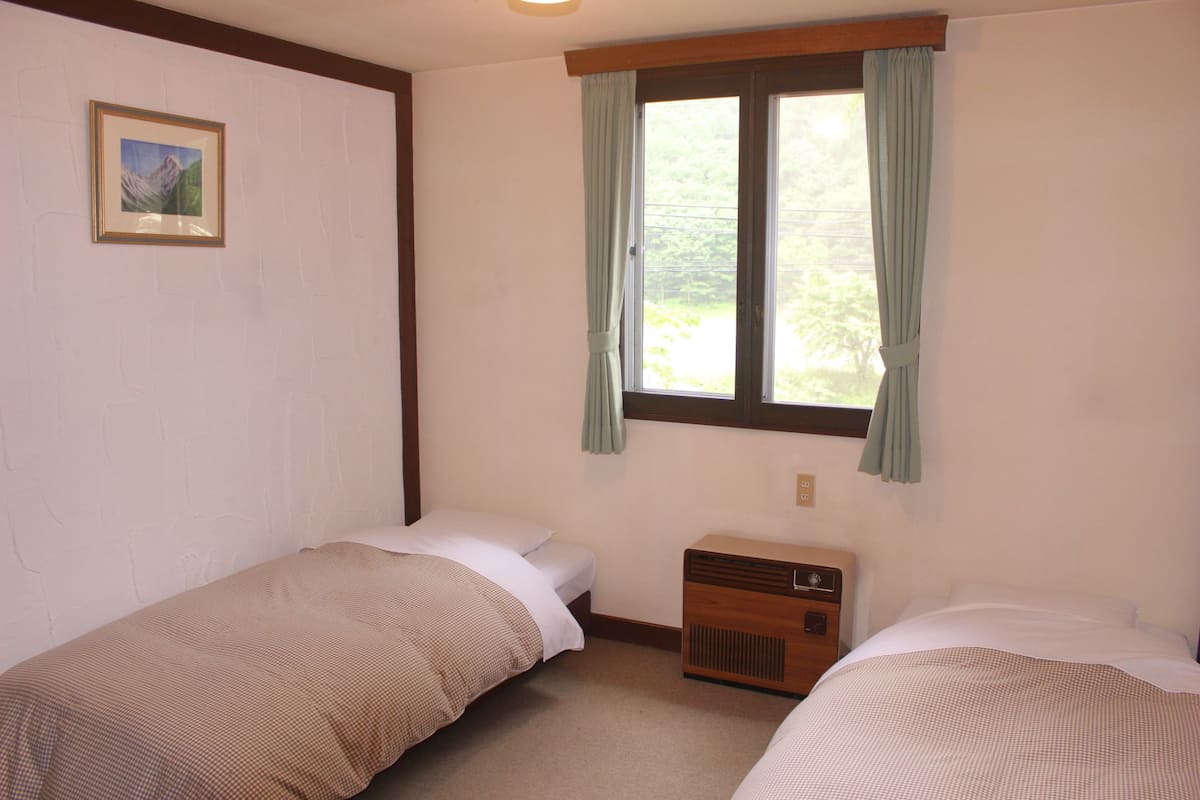 Una habitación con dos camas individuales, una mesita de noche de madera, un calentador fijado a la pared y una ventana con cortinas.