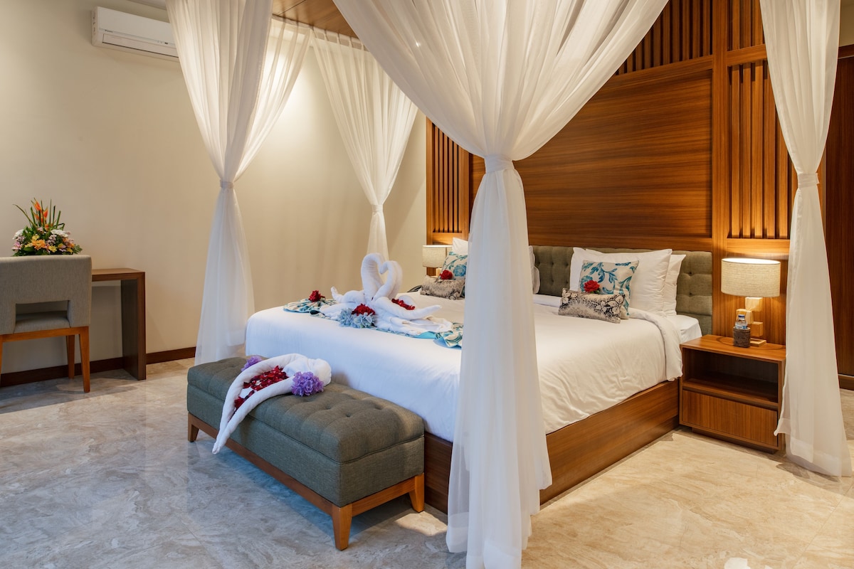 A bedroom with a canopy bed, a bedside table, a lamp, and a small stool.