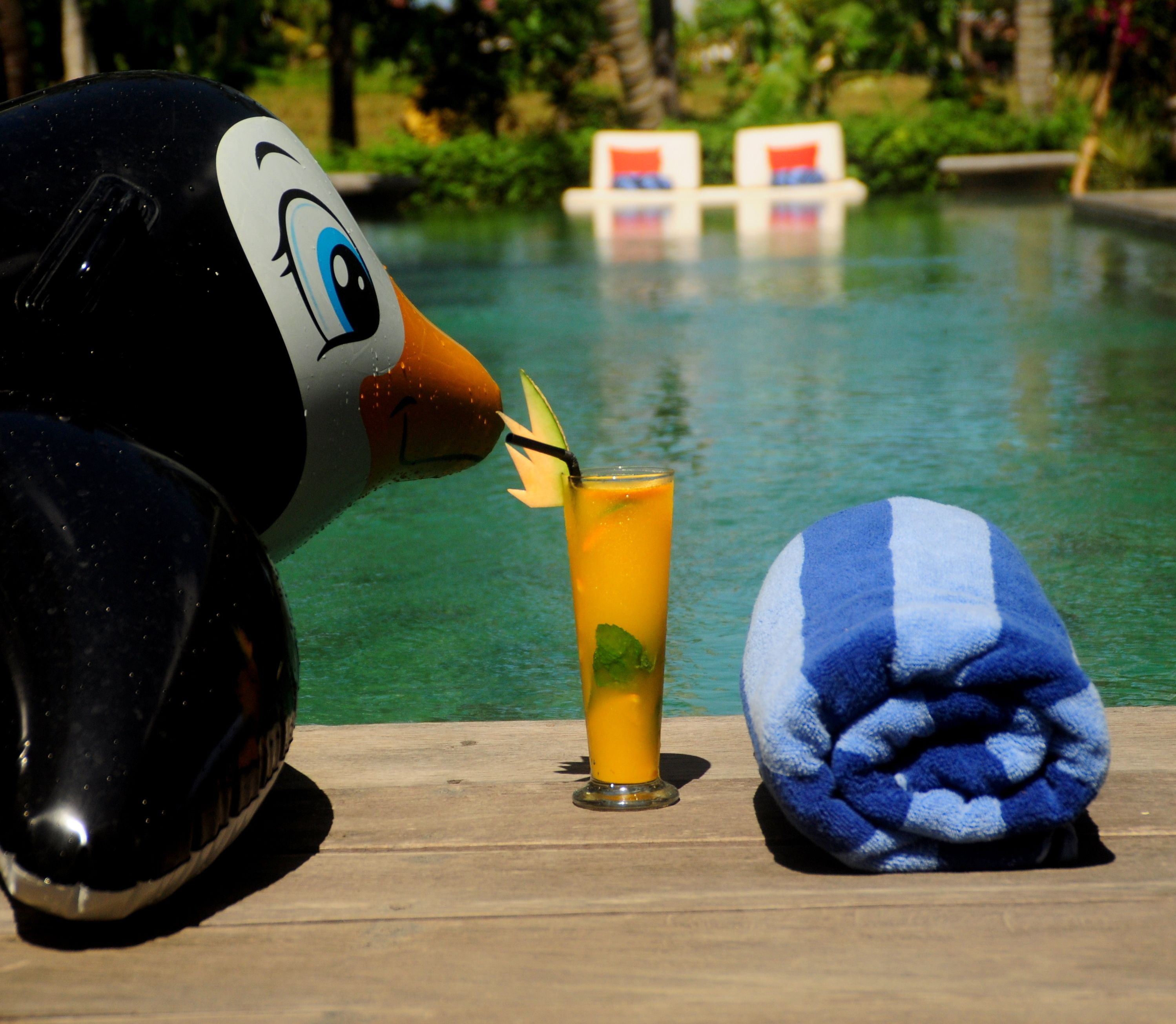 Um brinquedo de penguin macio, um copo com bebida amarela e uma toalha azul e branca sobre um deck de madeira perto de uma piscina.