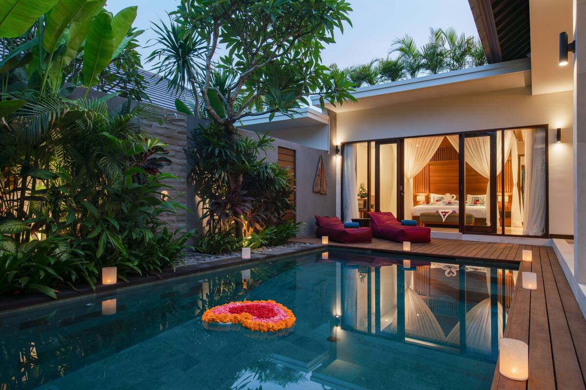 A modern outdoor pool area with a wooden deck, a glass-enclosed bedroom with a bed and curtains, and lush tropical plants.
