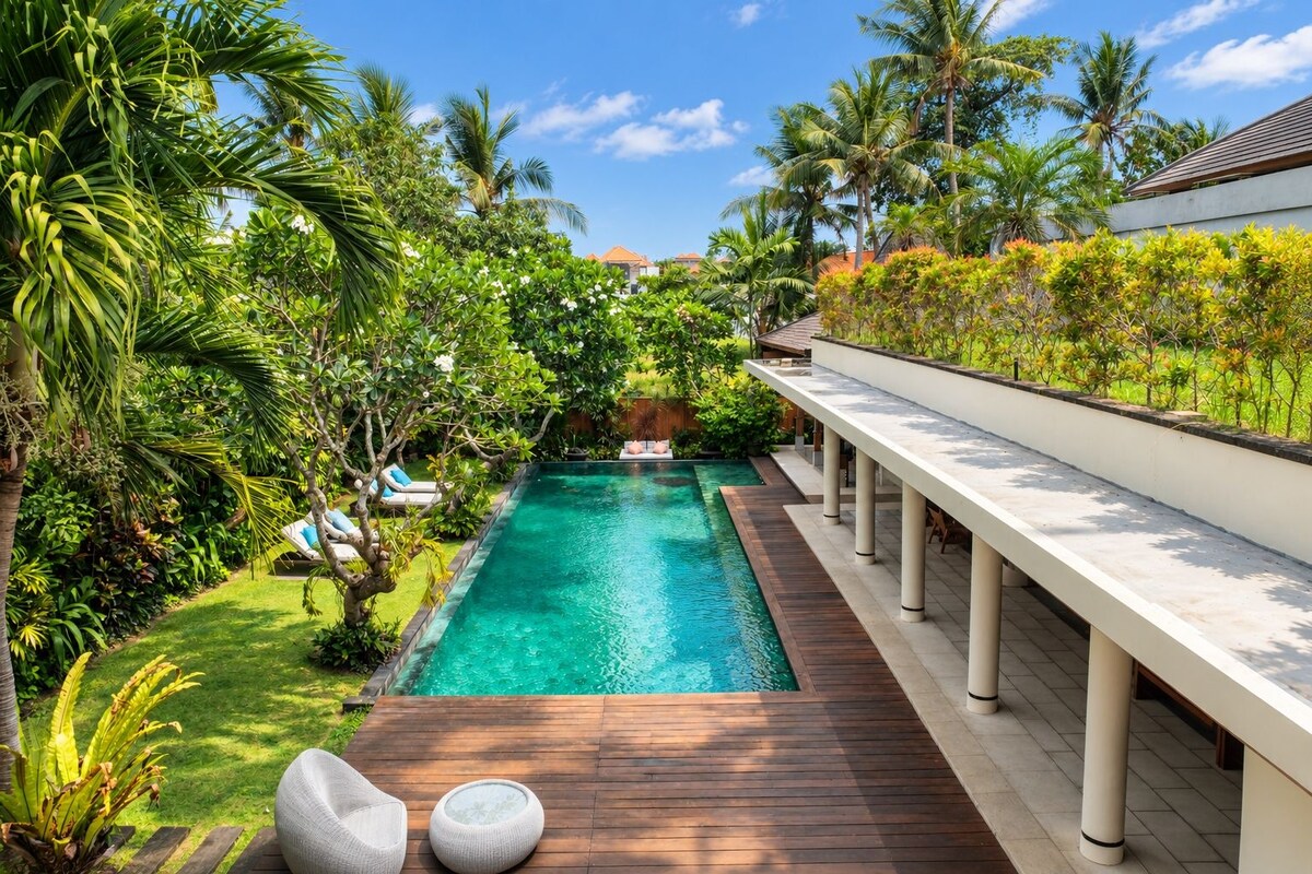 A swimming pool with a wooden deck, surrounded by lush greenery and palm trees.