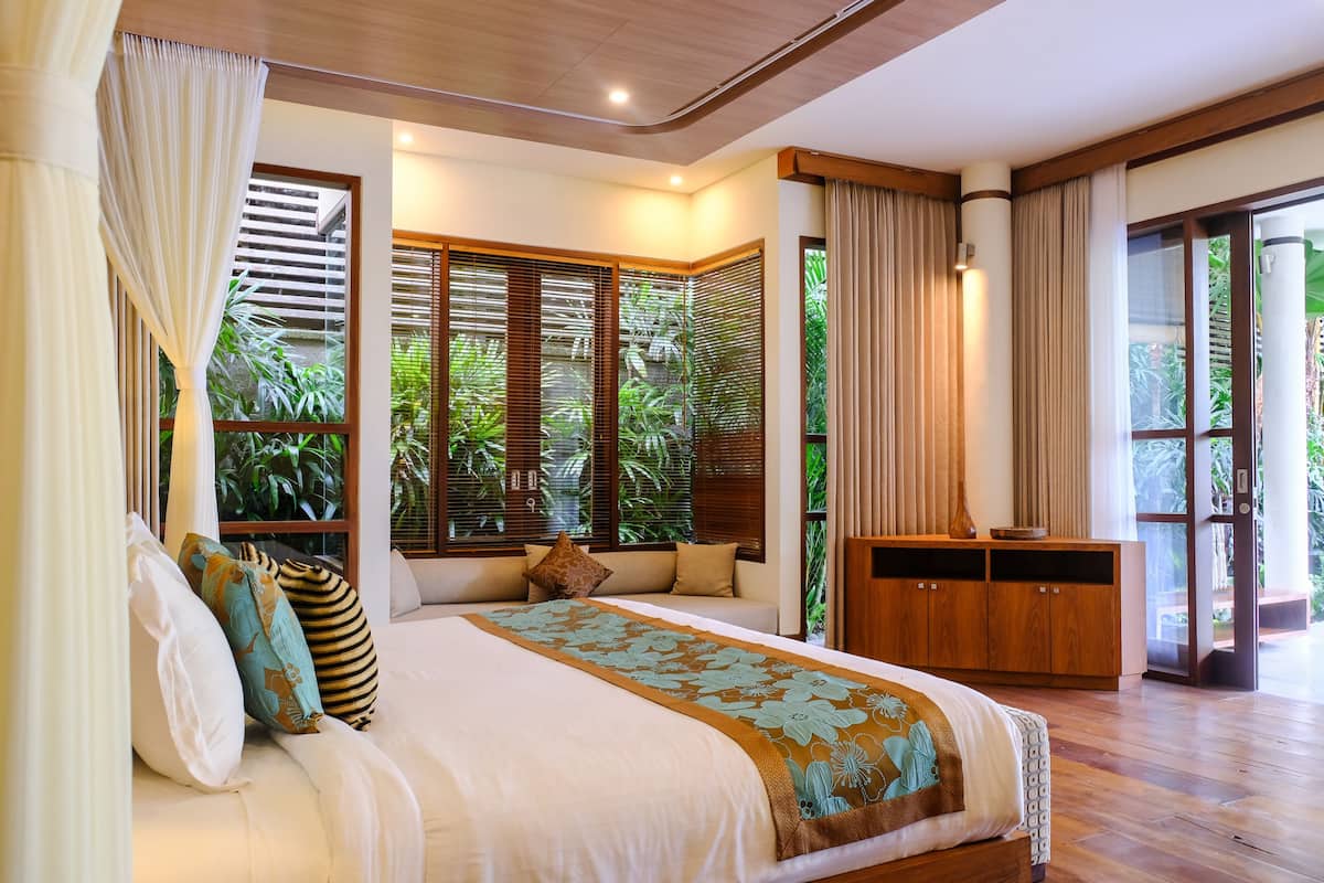 A bedroom with a large bed, wooden headboard, and a view of greenery through the windows.