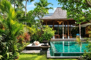 A poolside area with a covered seating area, lush greenery, and a clear blue pool.