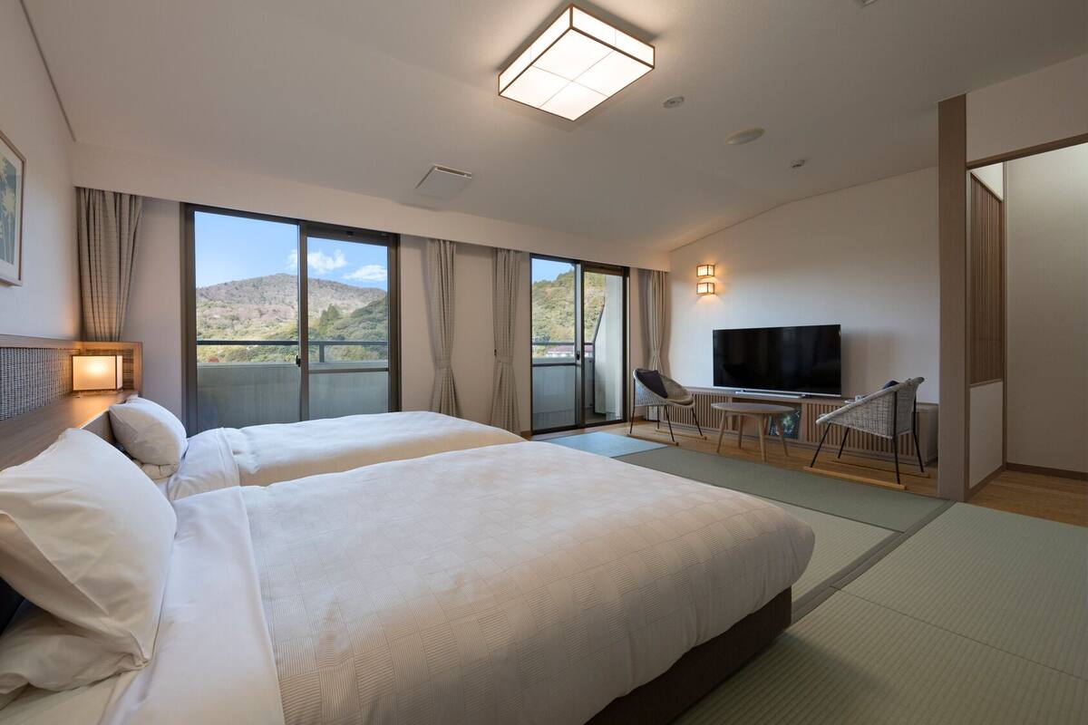 Habitación de hotel con dos camas, televisión, balcón con vista a las montañas y puerta corrediza.