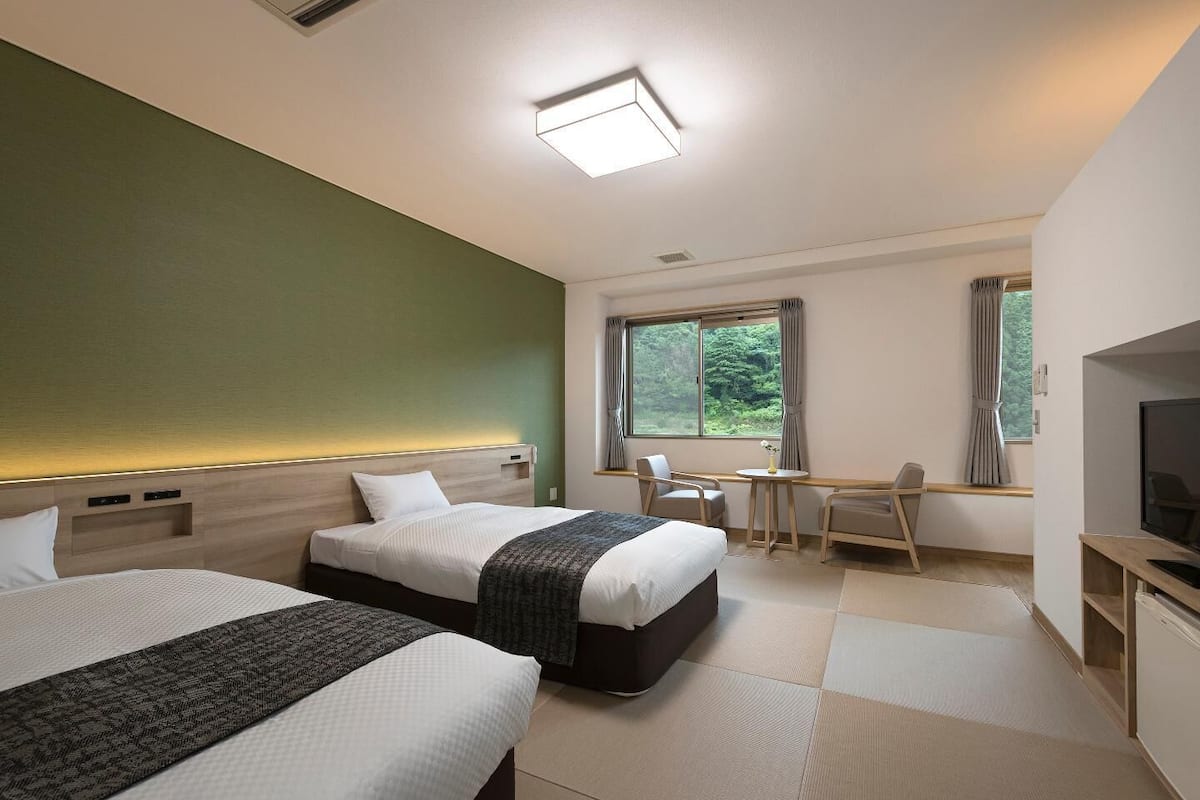 Habitación de hotel con dos camas, un escritorio, una silla, un televisor y una ventana con vistas a zonas verdes.