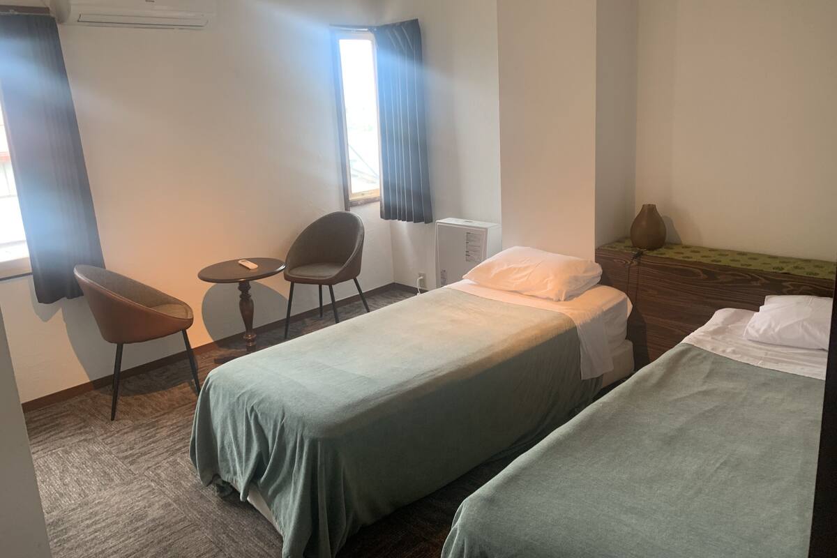 Ein Hotelzimmer mit zwei Betten, einem kleinen Tisch mit einem Stuhl, einem Fenster mit Vorhängen und einer Wandklimaanlage.