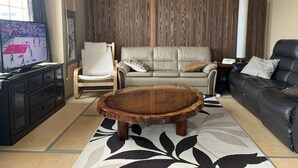 A living room with a wooden coffee table, a beige sofa, a black leather sofa, a wooden chair, and a television displaying a sports game. - Hakuba Ski Condos (Hakuba)