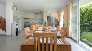A dining area with a wooden table set for six, surrounded by chairs, with a kitchen in the background and a view of greenery outside.