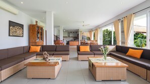 A spacious living area with two sofas, a coffee table, and a dining table. There is a kitchen in the background, and large windows provide natural light.
