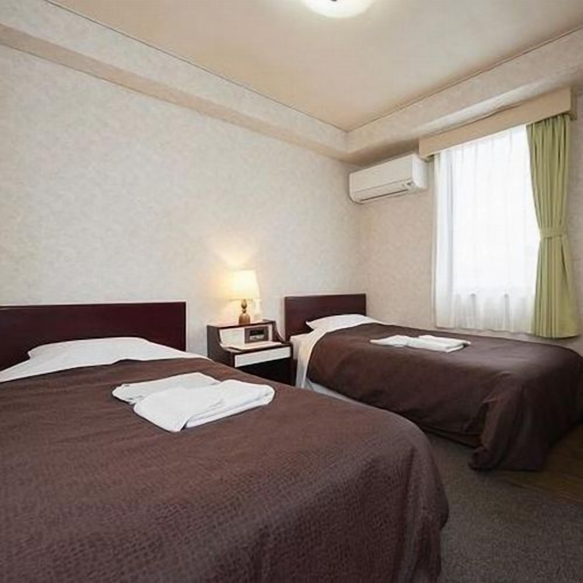 A hotel room with two beds, a nightstand with a lamp, an air conditioning unit, and a window with curtains.