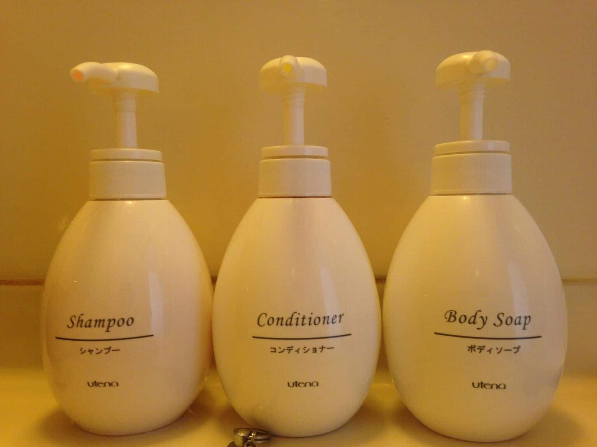 Drei Flaschen mit Körperpflegeprodukten, beschriftet mit 'Shampoo', 'Conditioner' und 'Duschseife', mit Pumpspendern, auf einer Oberfläche platziert.