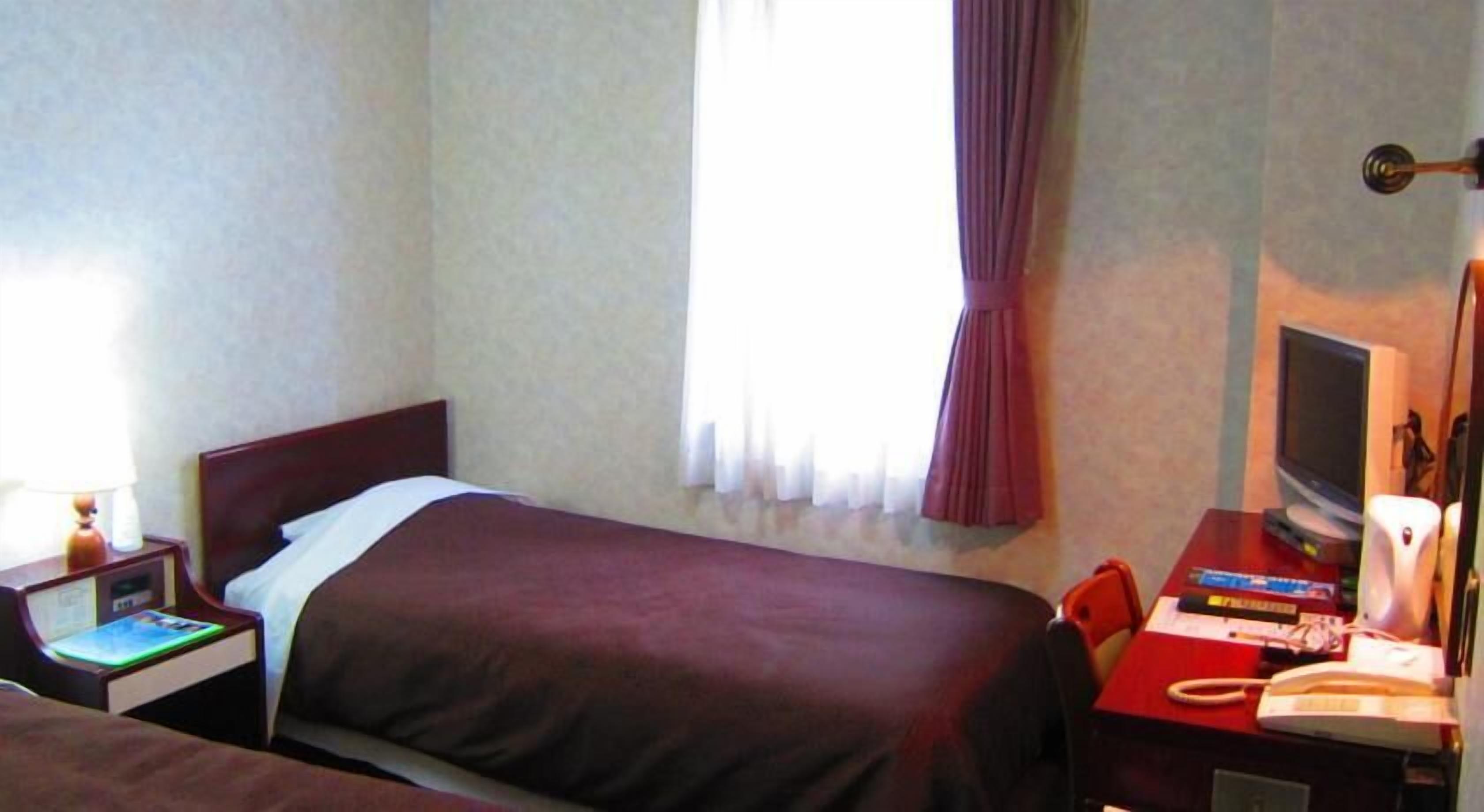 A hotel room with a single bed, a desk with a computer and a telephone, and a window with curtains.