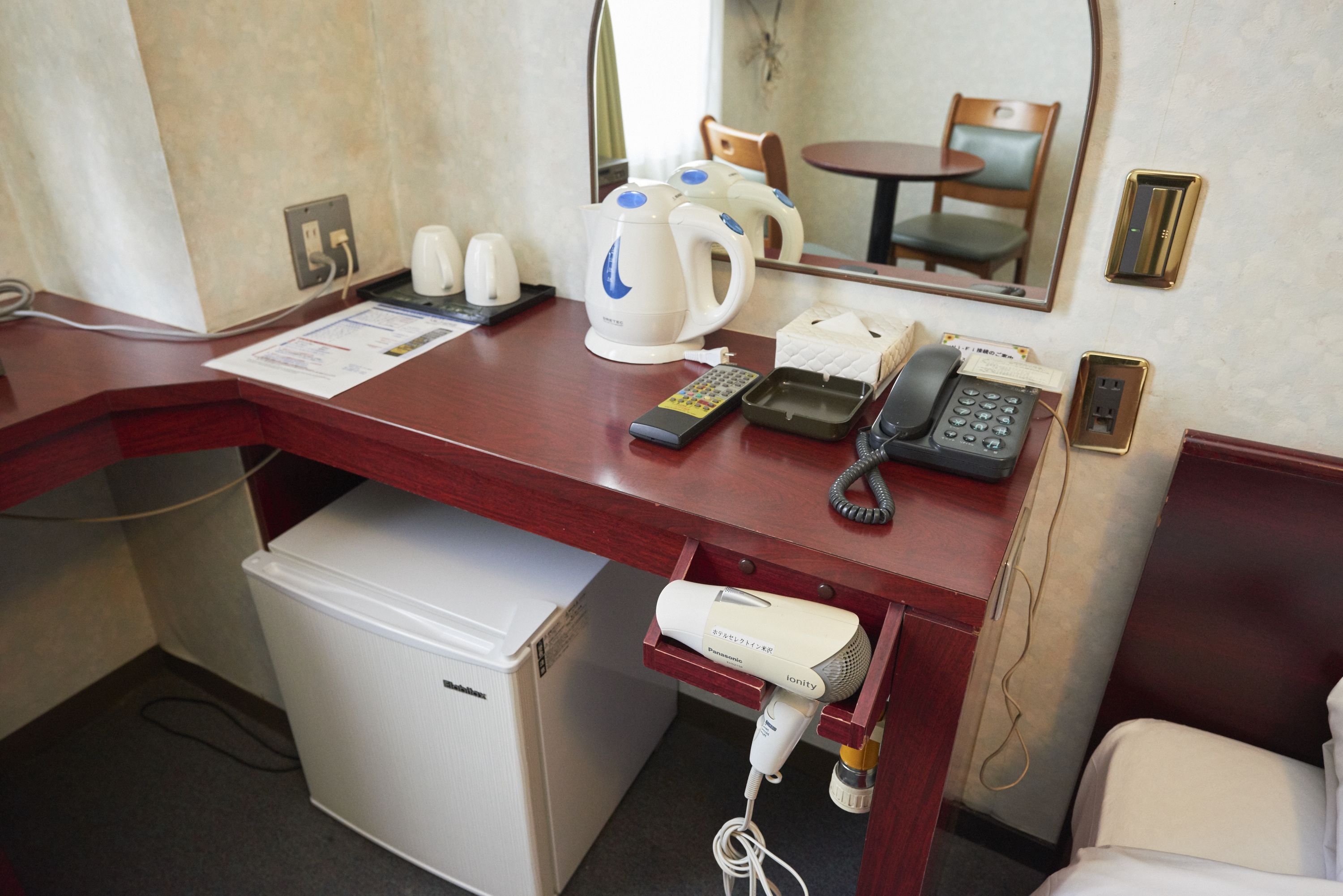 A hotel room desk with a refrigerator, a hairdryer, a telephone, a kettle, and a mirror.