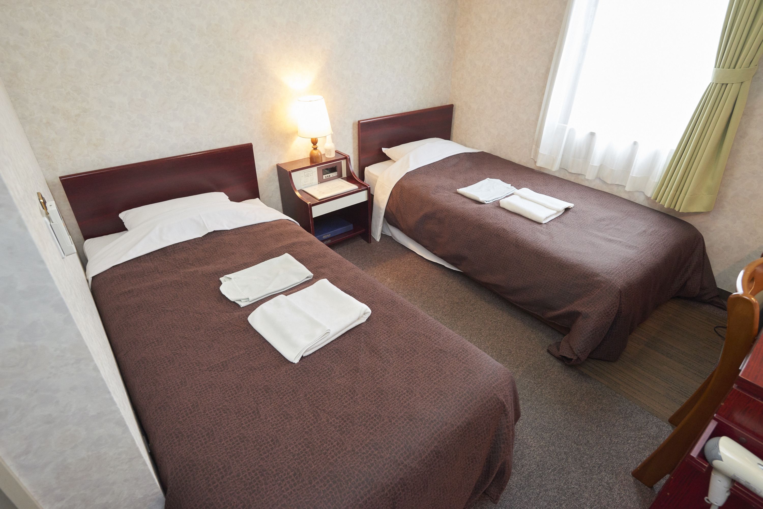 A hotel room with two beds, each with a brown bedspread and white pillows. There is a nightstand with a lamp between the beds, and a chair is visible in the corner.