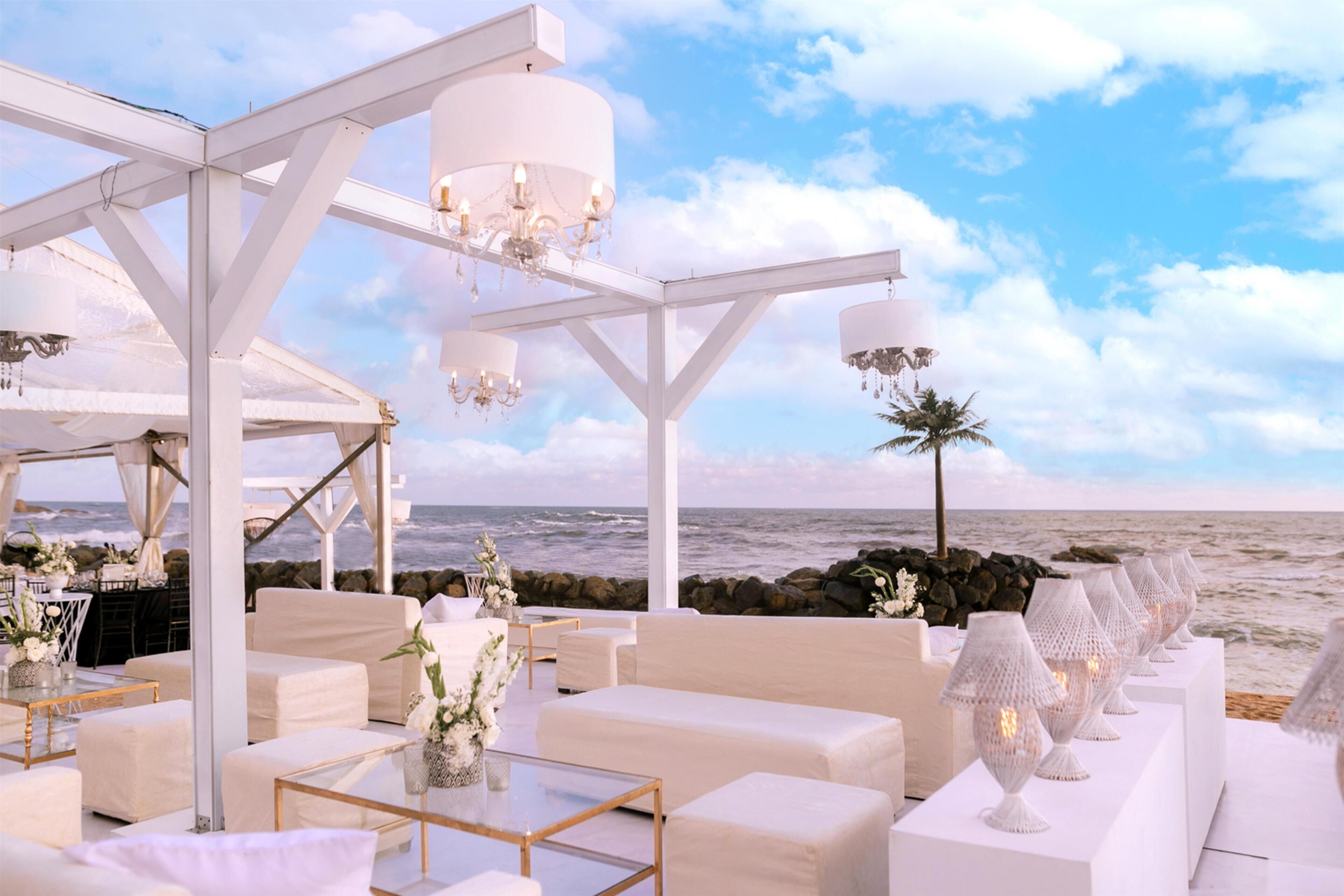 Un área de estar al aire libre con mobiliario blanco, candelabros y vista al océano.