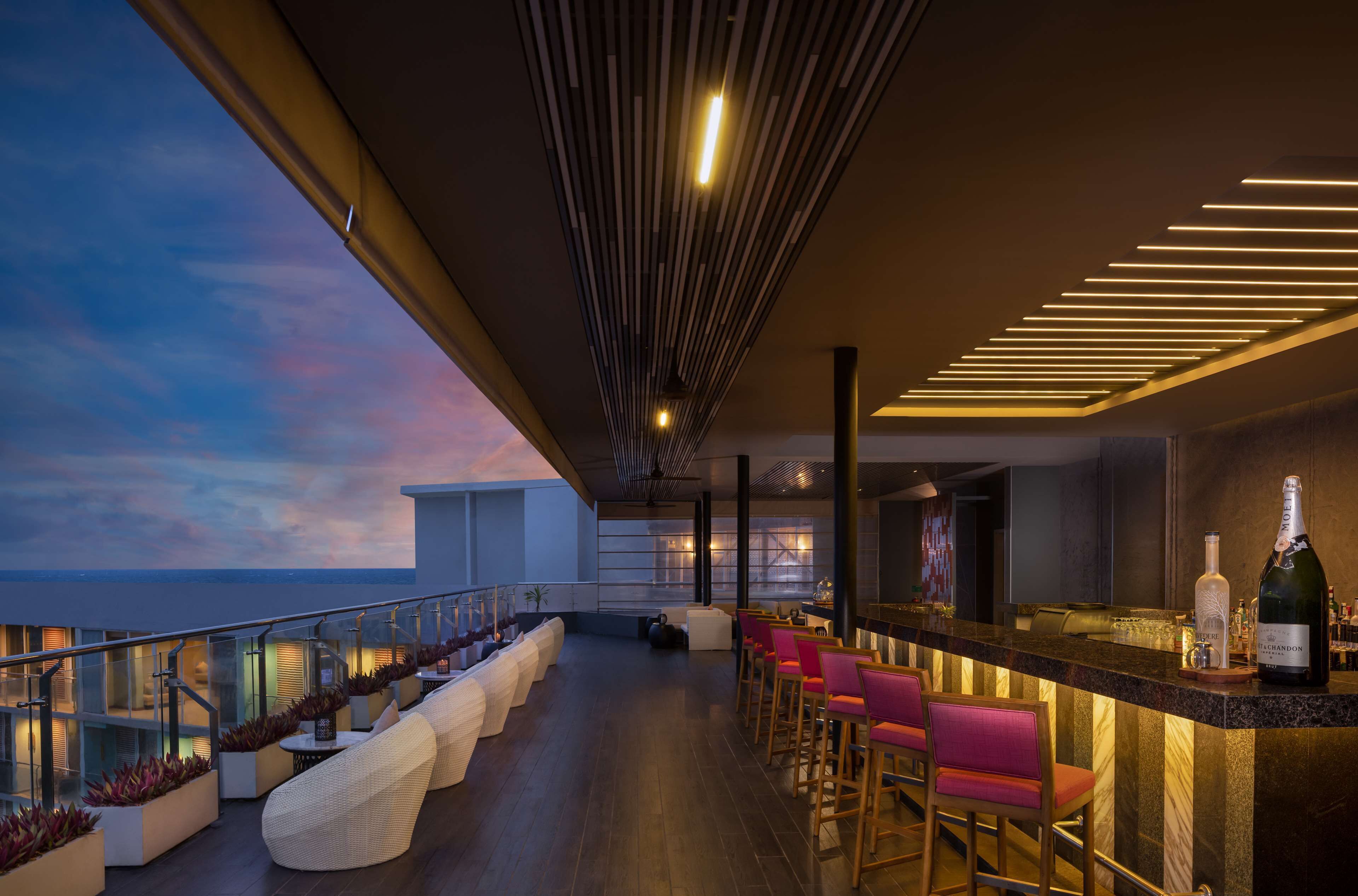 Eine moderne Bar mit einer langen Theke, Barhocker und einer Champagnerflasche. Gepolsterte Sitzbereiche und Blick zum Himmel.