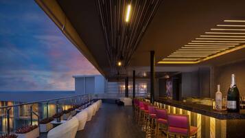 Eine moderne Bar mit einer langen Theke, Barhocker und einer Champagnerflasche. Gepolsterte Sitzbereiche und Blick zum Himmel.
