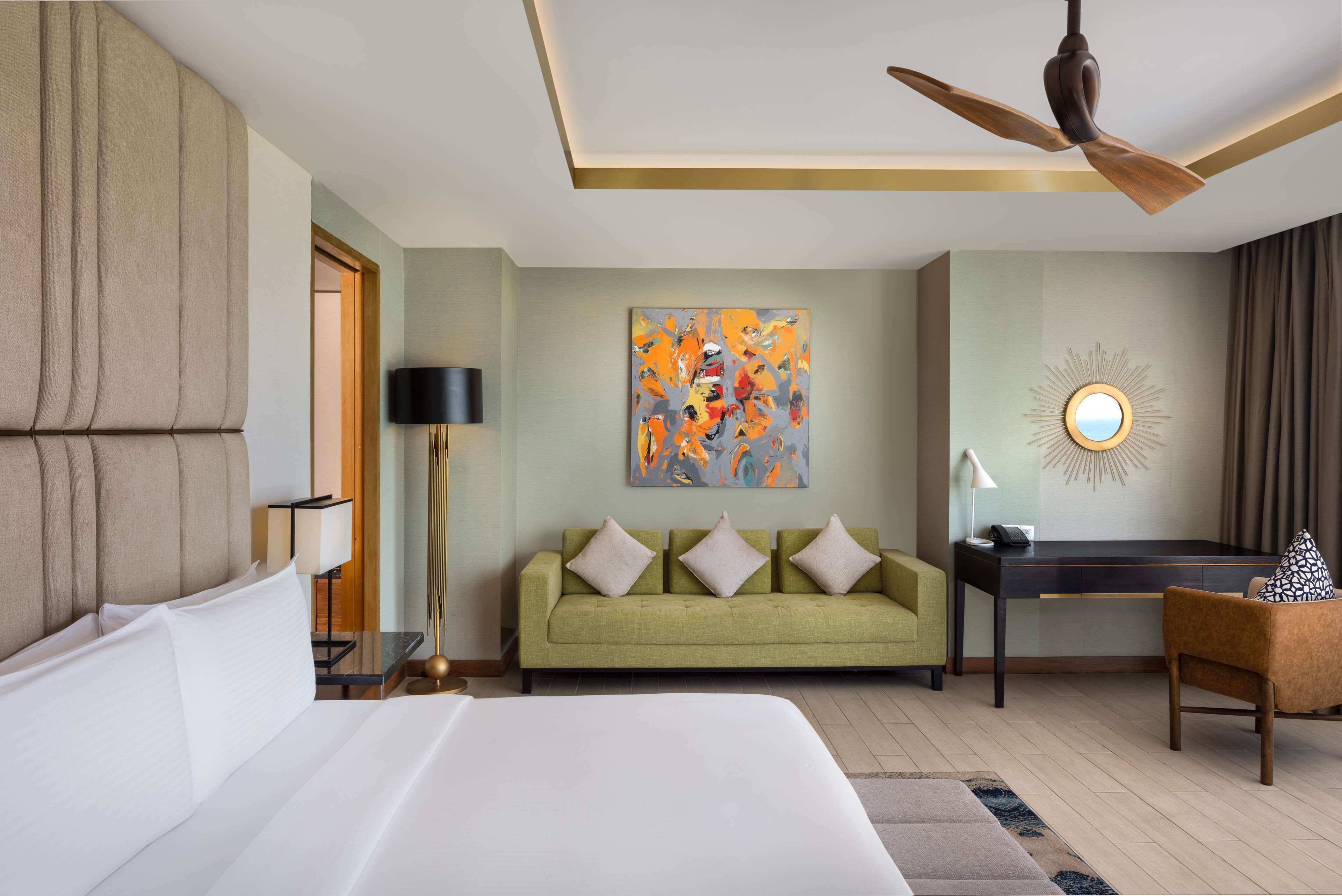 A hotel room with a bed, a green sofa, a desk, a ceiling fan, and a wall with a colorful abstract painting.