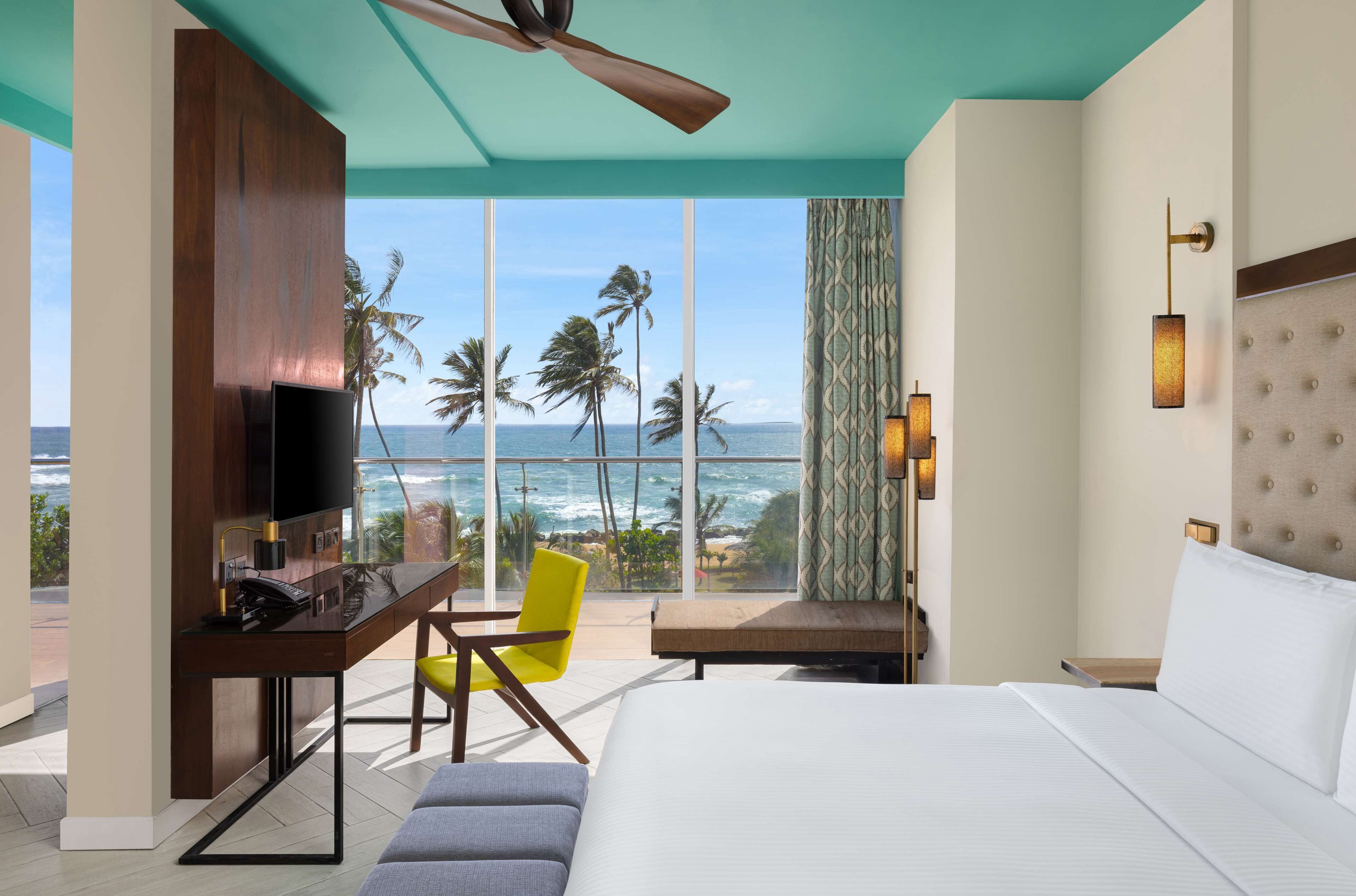 A hotel room with a large bed, a desk with a TV, a chair, and a view of the ocean.
