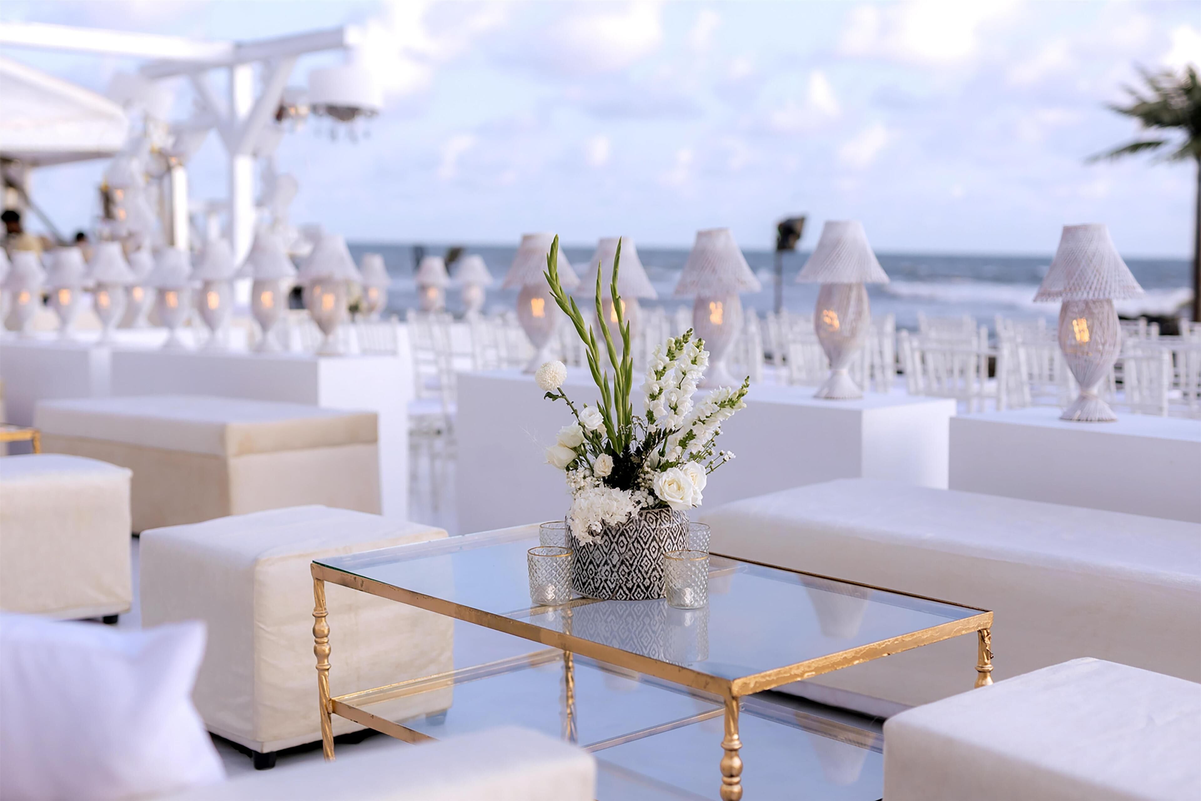 A beachside seating area with white furniture, a glass coffee table, and decorative lamps.