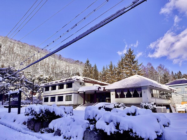 Un hotel con fachada nevada, zona de estacionamiento y montañas al fondo.