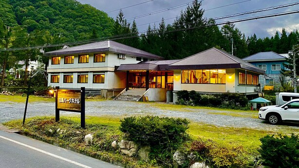 Un hôtel à deux étages, doté d’une enseigne, d’un espace extérieur abrité avec des sièges et offrant une vue sur un paysage montagneux.