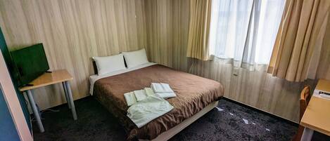 Quarto de hotel com cama, televisão, escrivaninha e janela com cortinas.