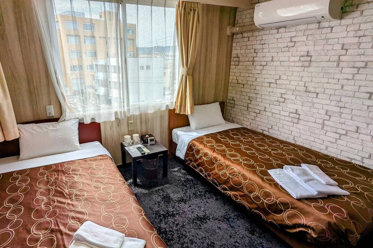 A hotel room with two beds, a small table with a radio, and a window with curtains.