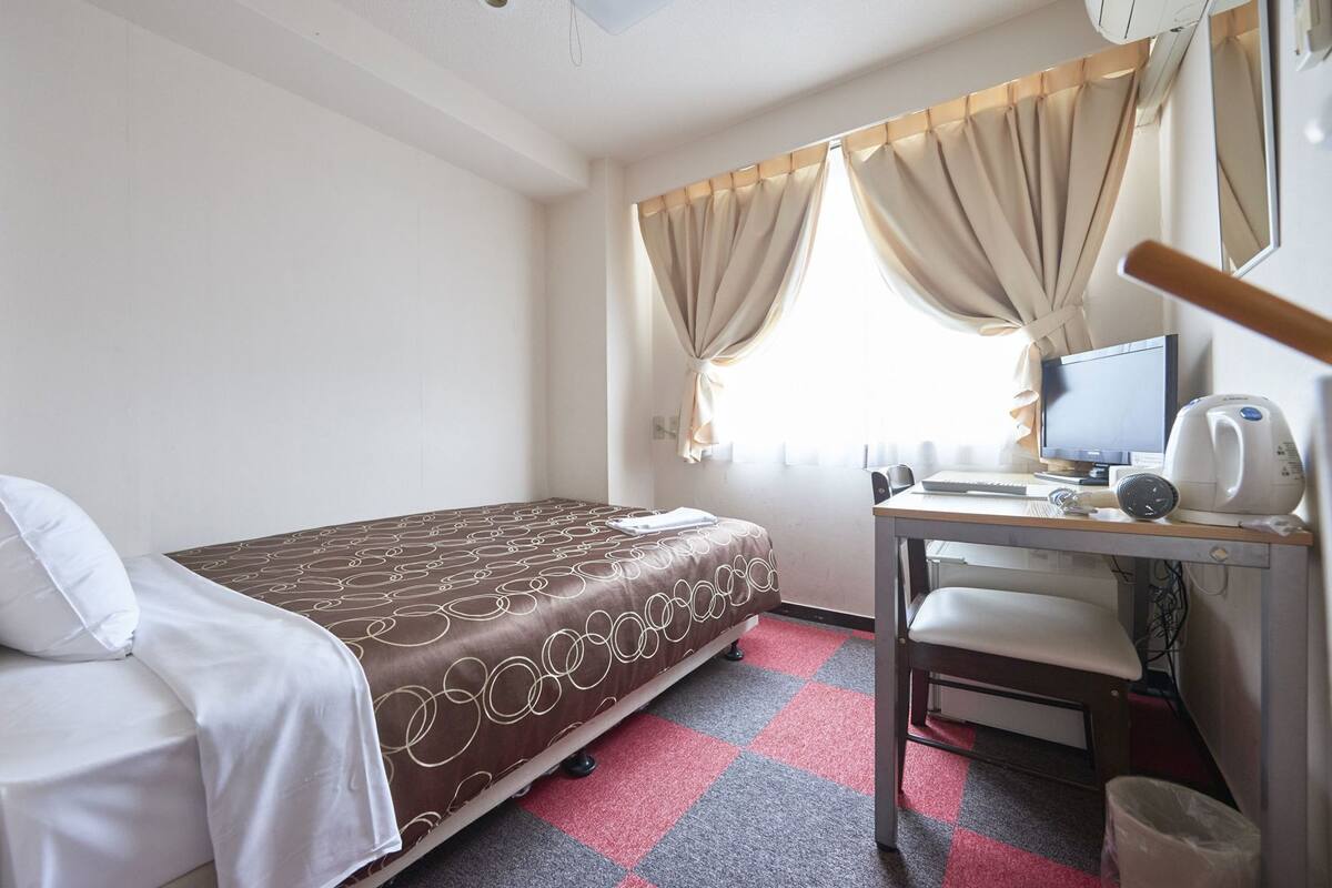 A hotel room with a bed, a desk with a computer, a chair, and a window with curtains.