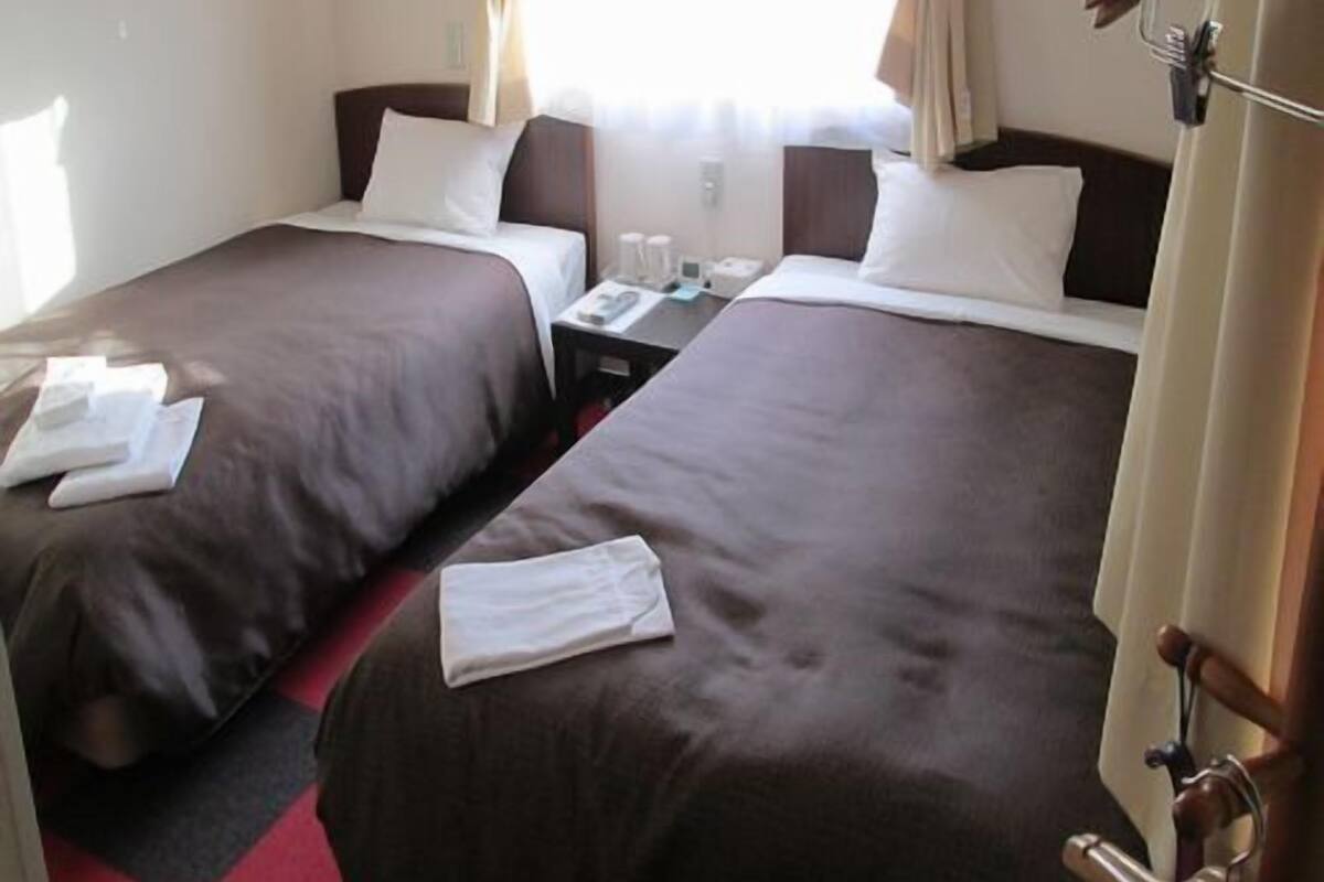 A hotel room with two beds, each with a pillow and a blanket. There is a nightstand with a lamp and a phone.