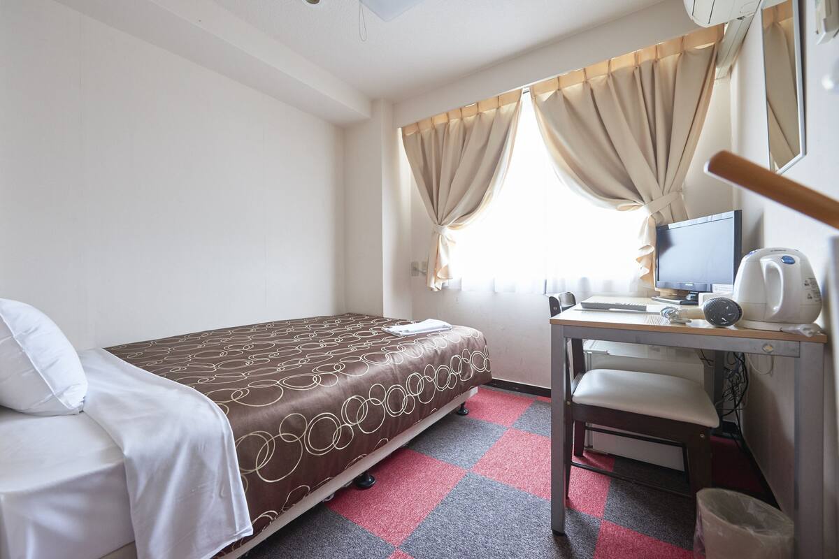 A hotel room with a bed, a desk with a computer, a chair, a window with curtains, and a trash bin.
