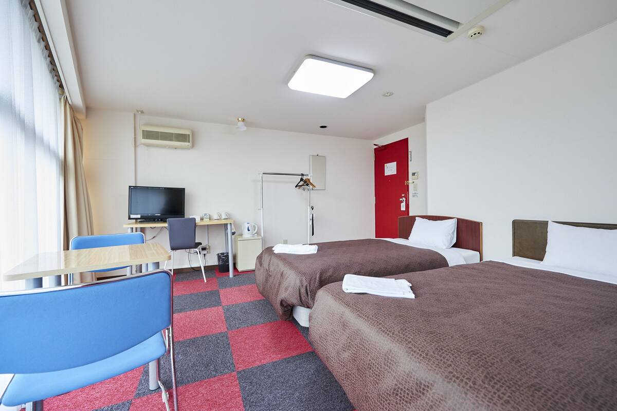 A hotel room with two beds, a desk, a chair, a television, and a red door.