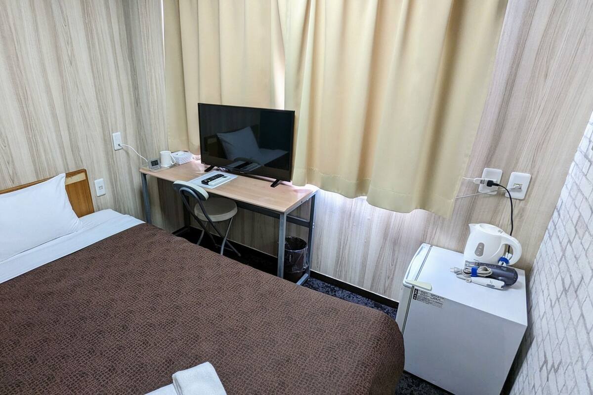A hotel room with a bed, a desk with a TV, a small kitchenette, and a window with curtains.