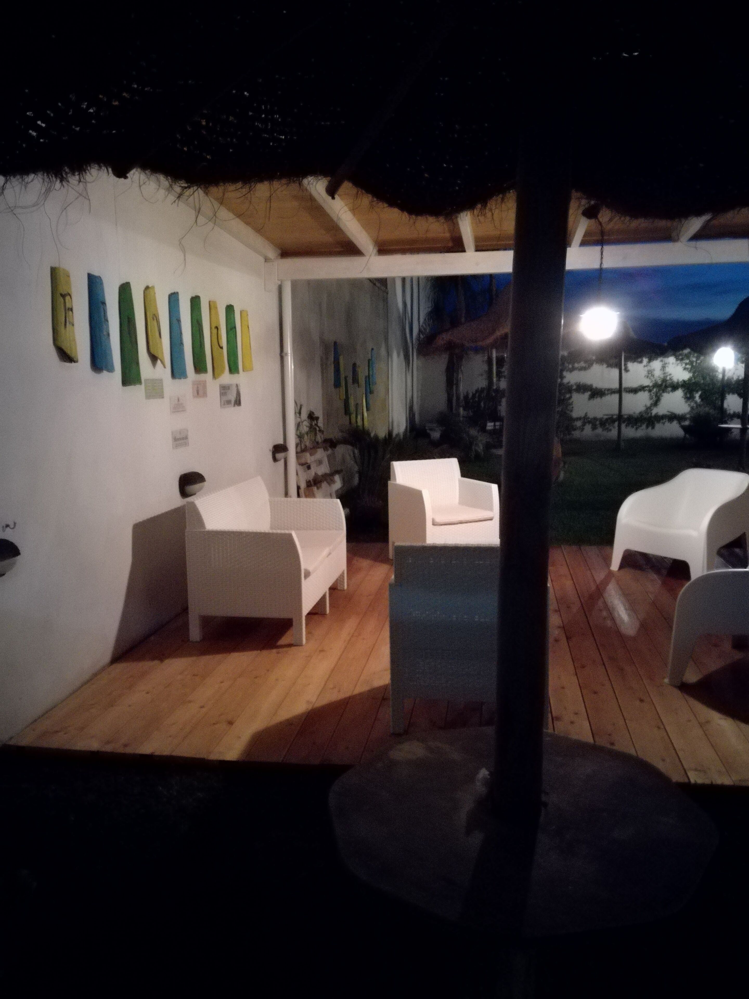 A covered outdoor seating area with white furniture, a wooden floor, and a wall with colorful artwork.