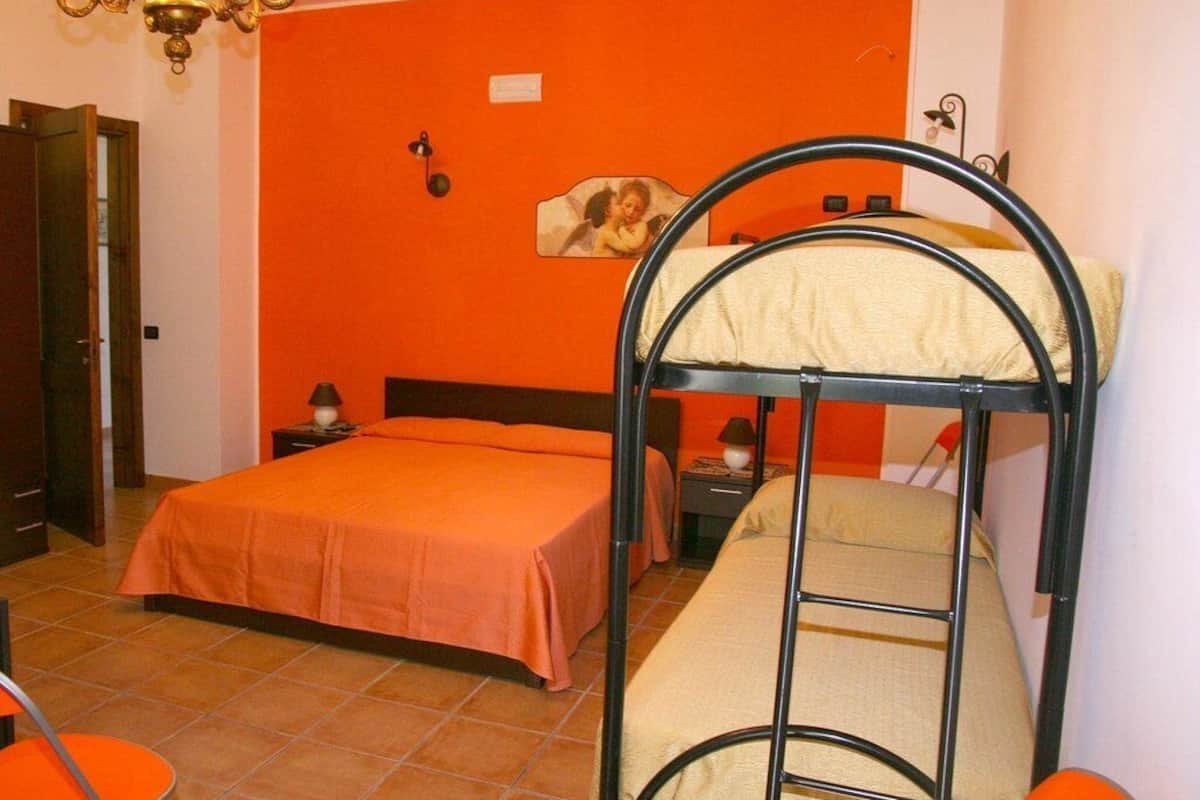 A room with two beds, one with a bunk and one single. There is a nightstand with a lamp, a picture on the wall, and a door to the left.