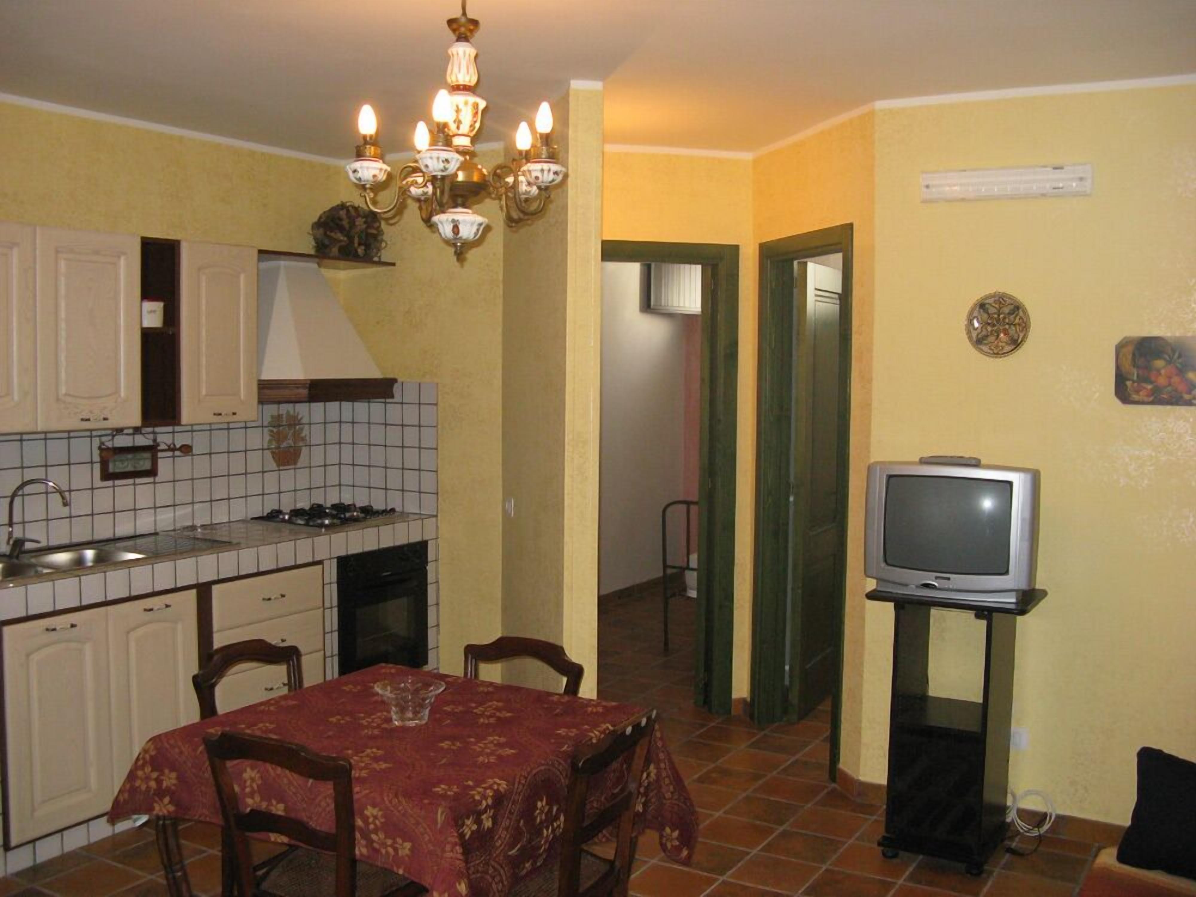 A compact kitchen with a tiled backsplash, a dining table with chairs, a chandelier, and a television on a stand.