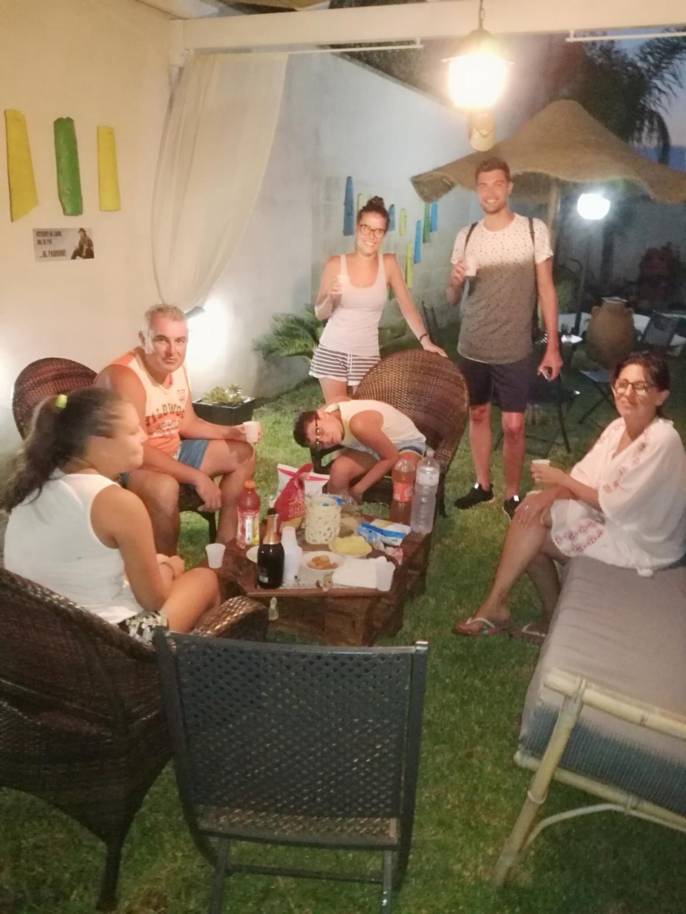 Un groupe de personnes est assis autour d’une table, où se trouvent de la nourriture et des boissons, dans un cadre extérieur agrémenté d’une tente et de chaises.