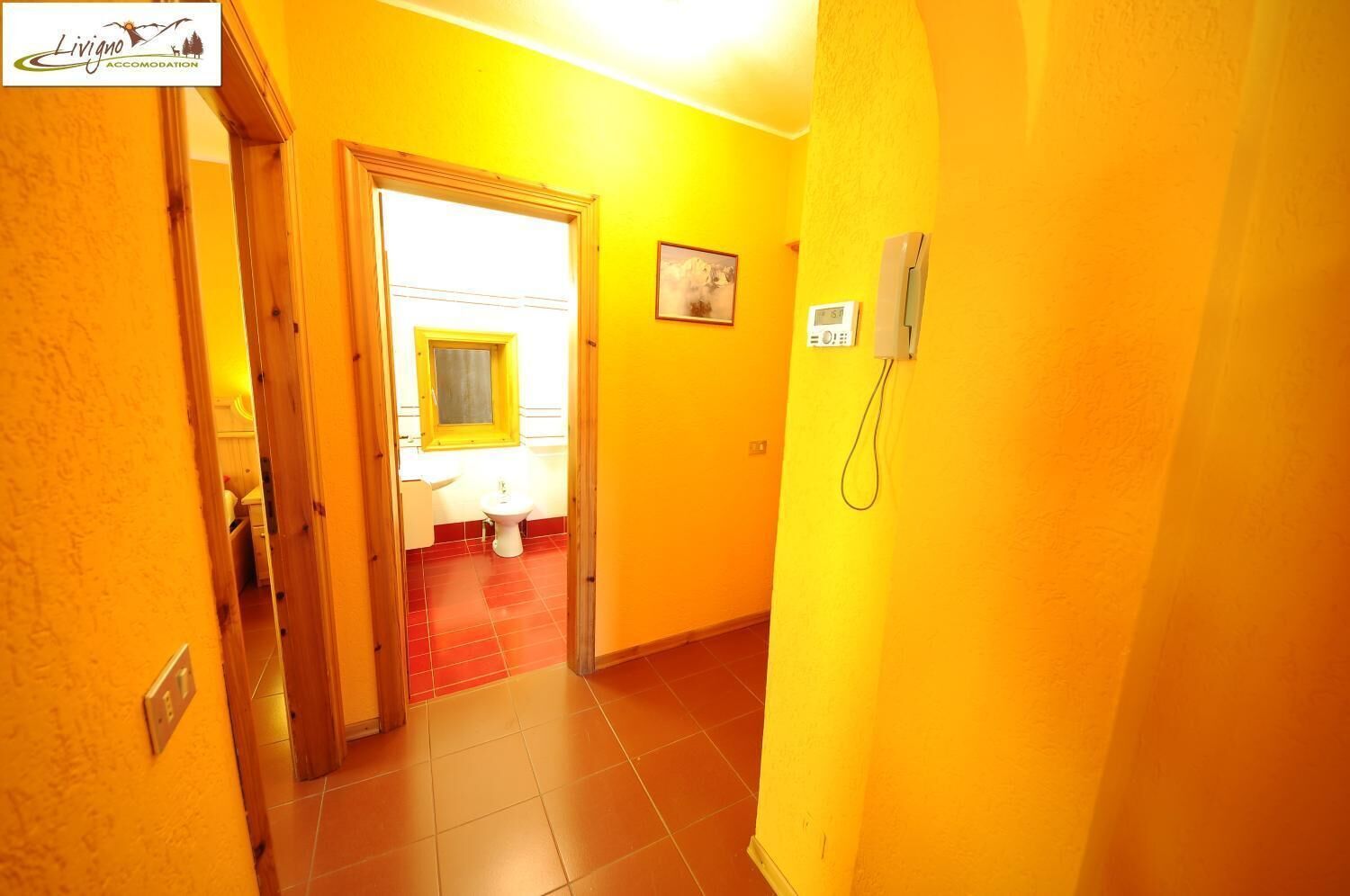 A narrow hallway with yellow walls and a red-tiled floor leading to a bathroom with a white sink and a yellow-framed window.