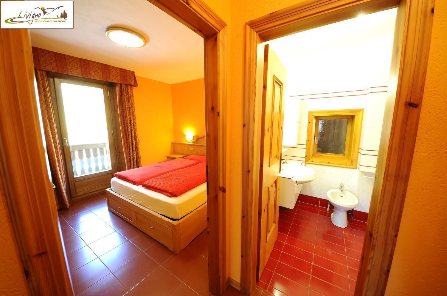 A bedroom with a bed, a bedside table, a window with curtains, and a bathroom with a sink and mirror.