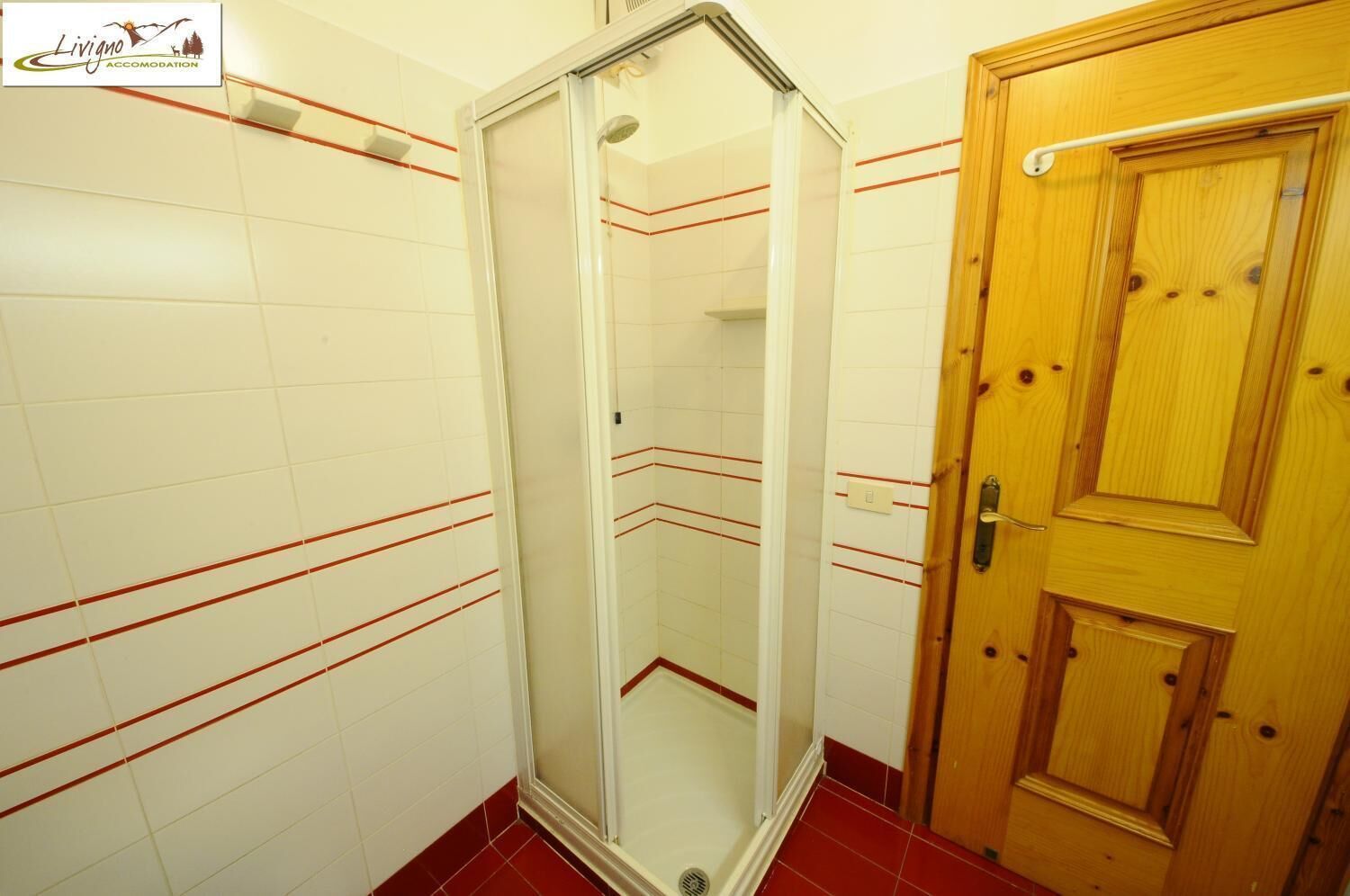 Un baño con paredes de azulejos blancos, cabina de ducha con mampara de vidrio y puerta de madera.